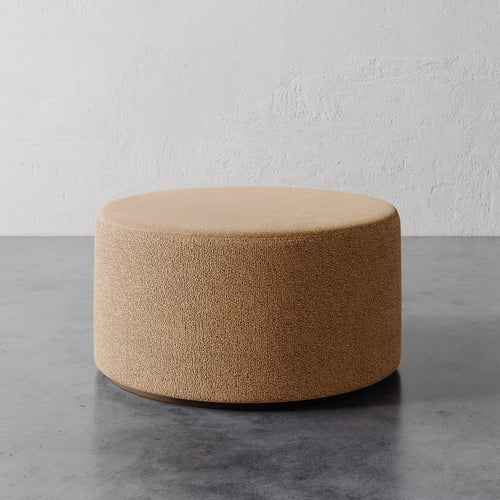 Lemieux et Cie Sophie Round 36" Ottoman in warm textured upholstery, cylindrical form on concrete floor against textured plaster wall.