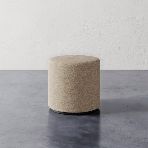 Lemieux et Cie Sophie Round Ottoman in warm neutral upholstery with dark base, cylindrical pouf shown on concrete floor against textured plaster wall.