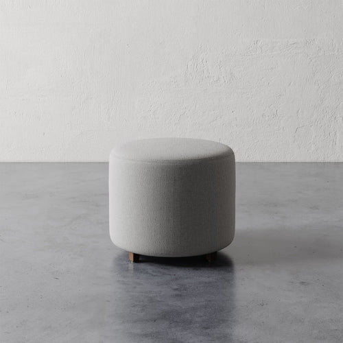 Lemieux et Cie Fabien 22" Round Ottoman, upholstered in warm gray, low cylindrical form on concrete floor against textured plaster wall.