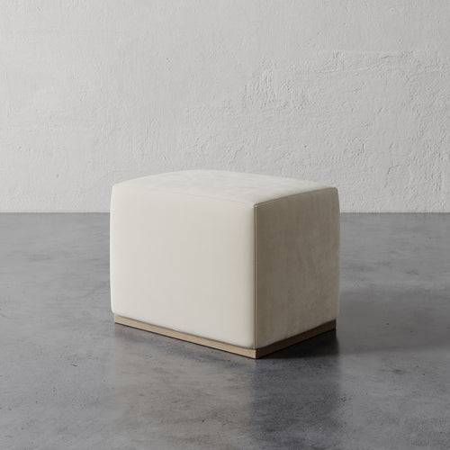 Lemieux et Cie Sophie Rectangular Ottoman in ivory upholstery with taupe side panel, low-profile ottoman on stone flooring against textured plaster walls.