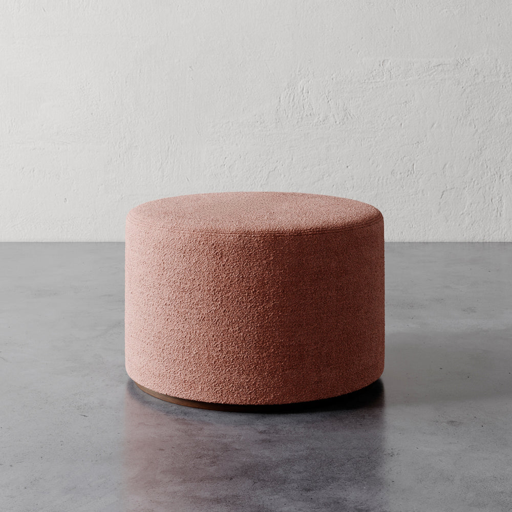 Lemieux et Cie Sophie Round Ottoman in blush bouclé, cylindrical upholstered ottoman on polished concrete floor against textured plaster wall.