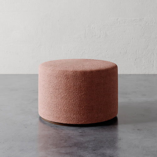 Lemieux et Cie Sophie Round Ottoman in blush bouclé, cylindrical upholstered ottoman on polished concrete floor against textured plaster wall.