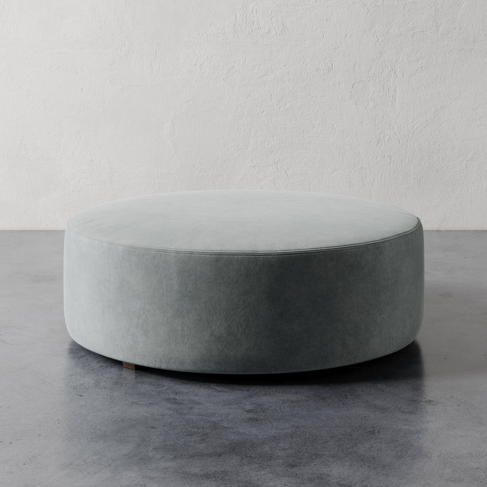 Lemieux et Cie Fabien 48" Round Ottoman, large-scale upholstered ottoman in pale sage, shown on polished concrete floor against textured white plaster walls.
