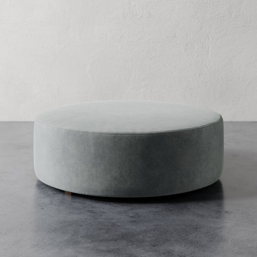 Lemieux et Cie Fabien 48" Round Ottoman, large-scale upholstered ottoman in pale sage, shown on polished concrete floor against textured white plaster walls.