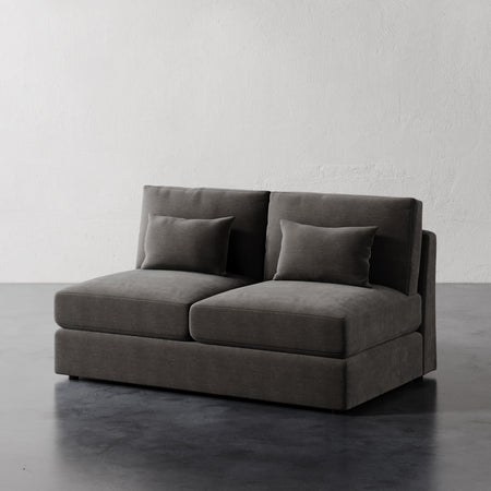 Lemieux et Cie Lacroix Armless Loveseat in charcoal gray, modular two-seat sofa with low profile and bolster pillows, on dark stone floor against textured plaster walls.
