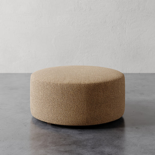 Lemieux et Cie Fabien 36" Round Ottoman, low-profile cylindrical ottoman in warm textured upholstery, shown on gray concrete floor against white plaster wall.