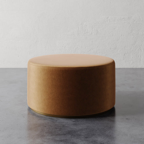 Lemieux et Cie Sophie Round Ottoman in warm caramel upholstery, low cylindrical form on concrete floor against textured plaster wall.