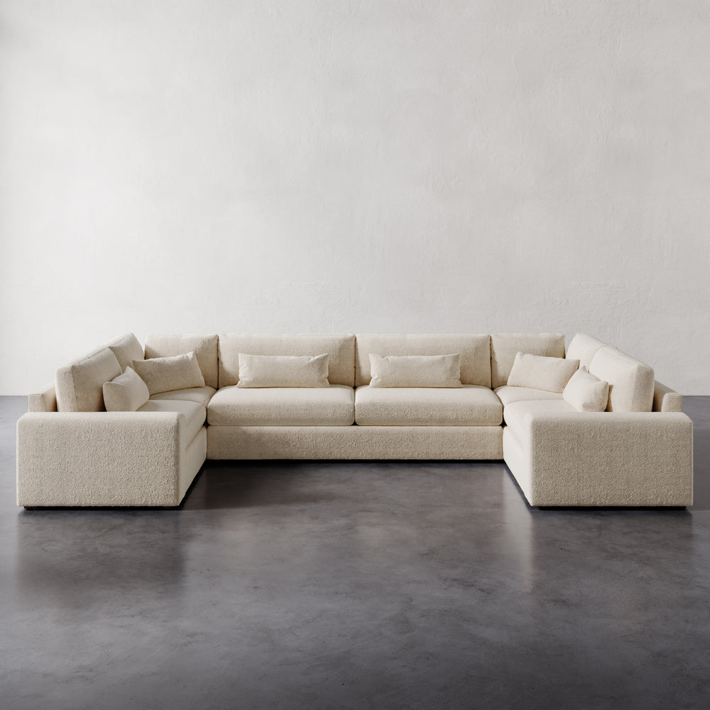 Lemieux et Cie Lacroix U-Sectional in ivory bouclé, modular sofa with deep cushions and bolster pillows, on dark concrete floors against neutral plaster walls.