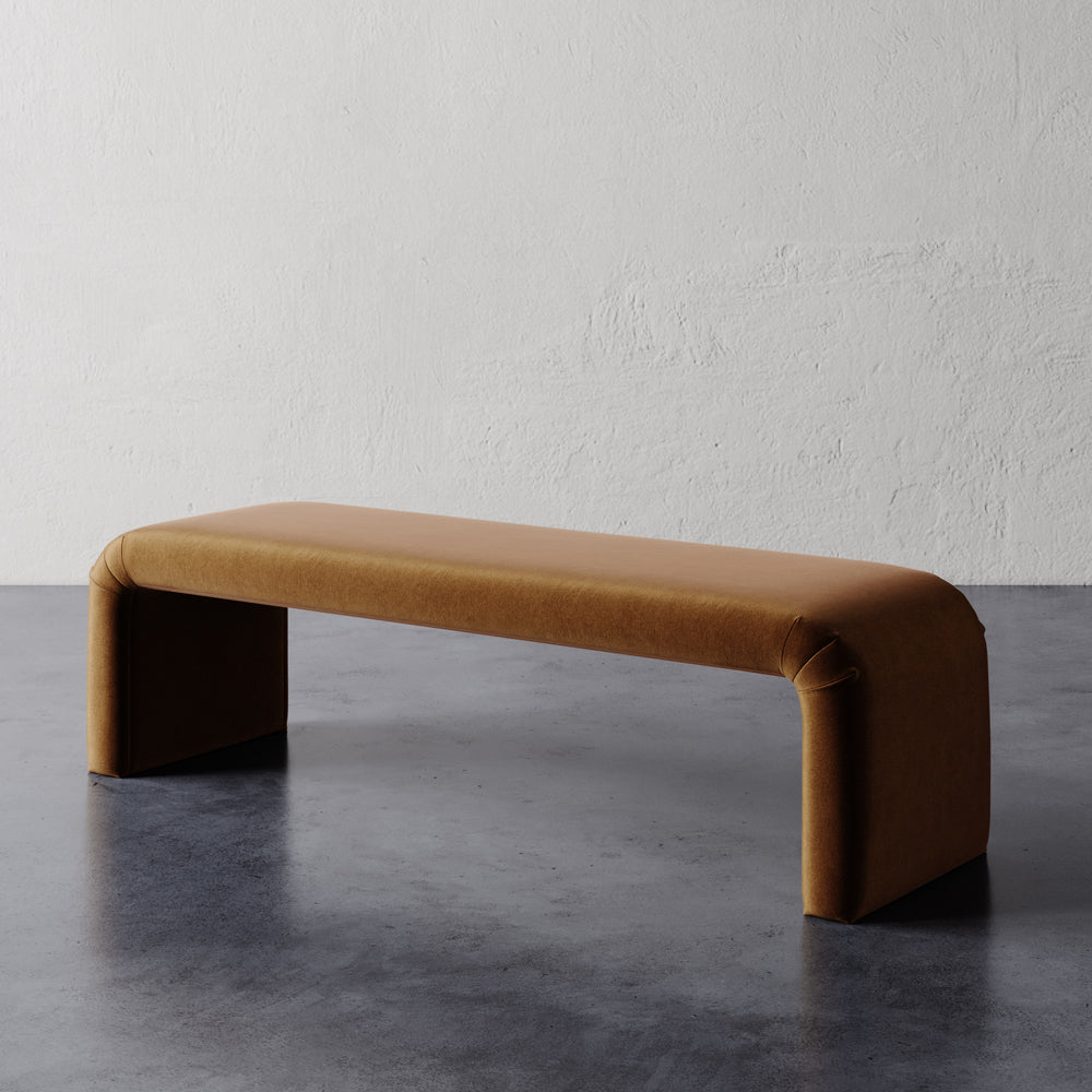 Lemieux et Cie Nadia 62" Bench in warm caramel, upholstered bench with rounded waterfall edges, shown on concrete floor against textured plaster wall.