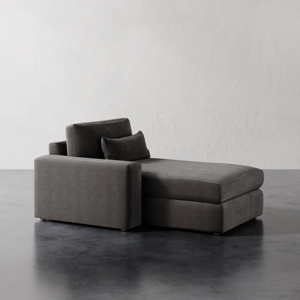 Lemieux et Cie Lacroix Left Chaise & A Half in charcoal, modular sectional with bolster pillow, on dark concrete floor against neutral plaster walls.