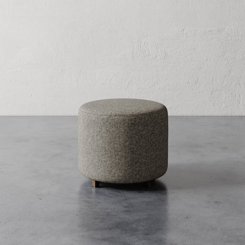 Lemieux et Cie Fabien 22" Round Ottoman in gray upholstery, low cylindrical ottoman on stone floor against textured plaster wall