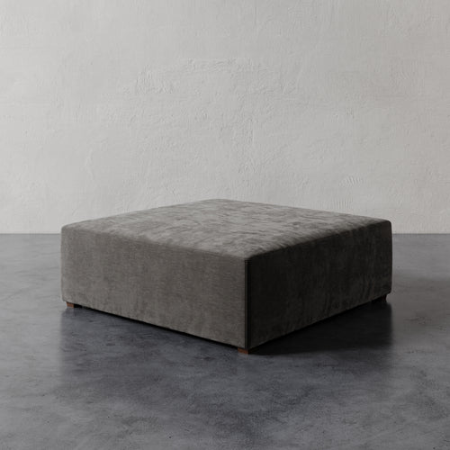 Lemieux et Cie Fabien 48" Square Ottoman in charcoal gray, low-profile upholstered ottoman on dark concrete floor against textured plaster wall.