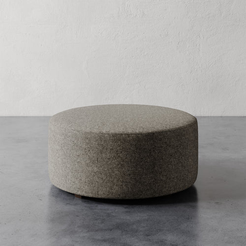 Lemieux et Cie Fabien 36" Round Ottoman in textured gray upholstery, low-profile ottoman on polished concrete floor against neutral plaster wall.