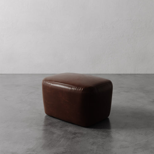 Lemieux et Cie Aurélie Leather Ottoman in warm brown leather, low-profile ottoman with rounded form, shown on concrete floor against textured plaster wall.