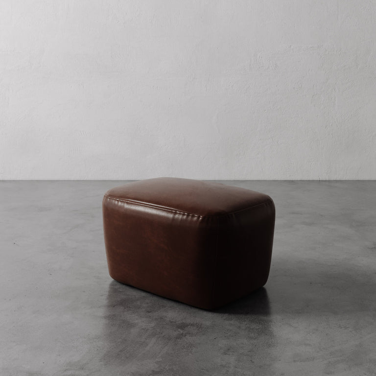 Lemieux et Cie Aurélie Leather Ottoman in warm brown leather, low-profile ottoman with rounded form, shown on concrete floor against textured plaster wall.