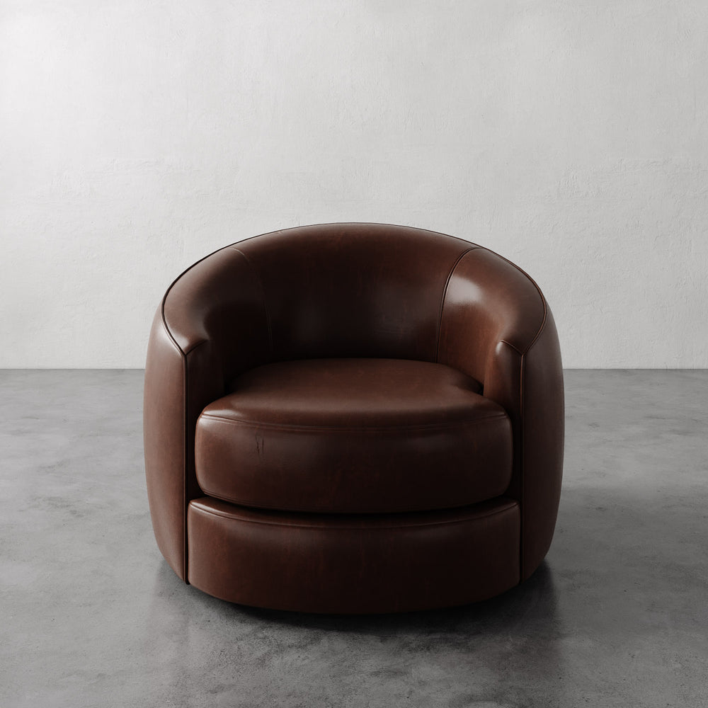 Lemieux et Cie Rulmann Swivel Chair in warm brown leather, rounded low-profile lounge chair on gray concrete floor with plaster walls.