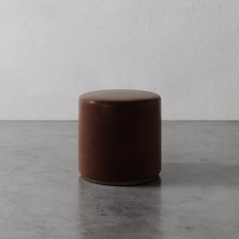 Lemieux et Cie Sophie Round Ottoman in warm tobacco brown leather, cylindrical form shown on concrete floor against textured plaster wall.