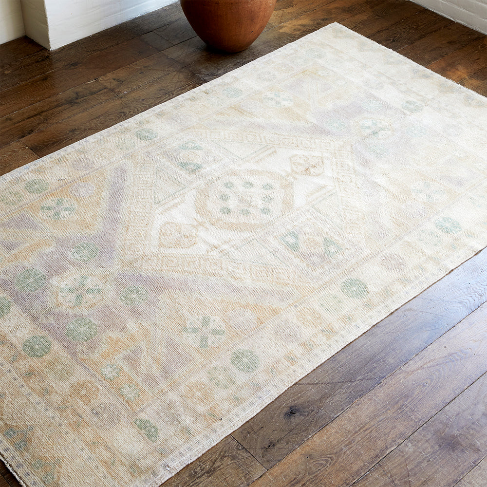 Lemieux et Cie Anatolian Village Rug in ivory with soft geometric motifs, vintage Turkish rug shown on weathered wood flooring beside terracotta vessel and white walls.