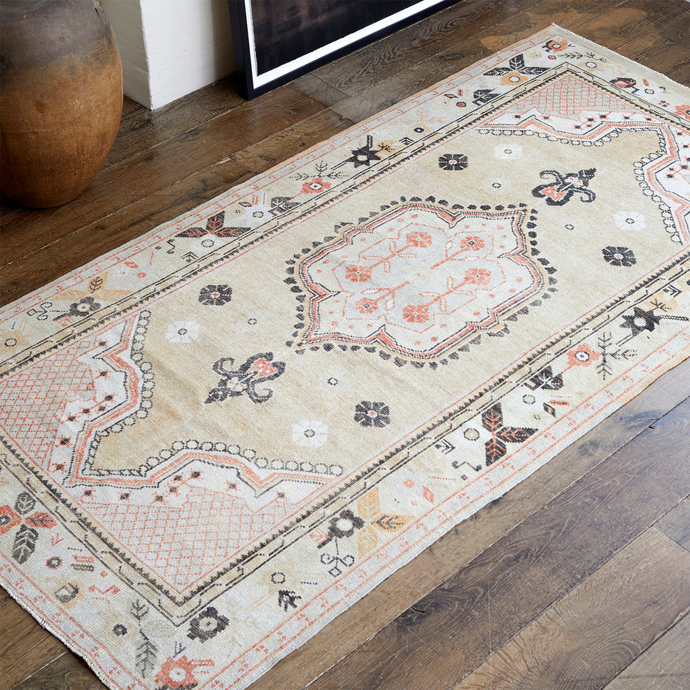 Lemieux et Cie Anatolian Village Rug in cream, coral and charcoal with geometric medallions, handwoven vintage wool rug on wood floors beside framed art and ceramic vessel.