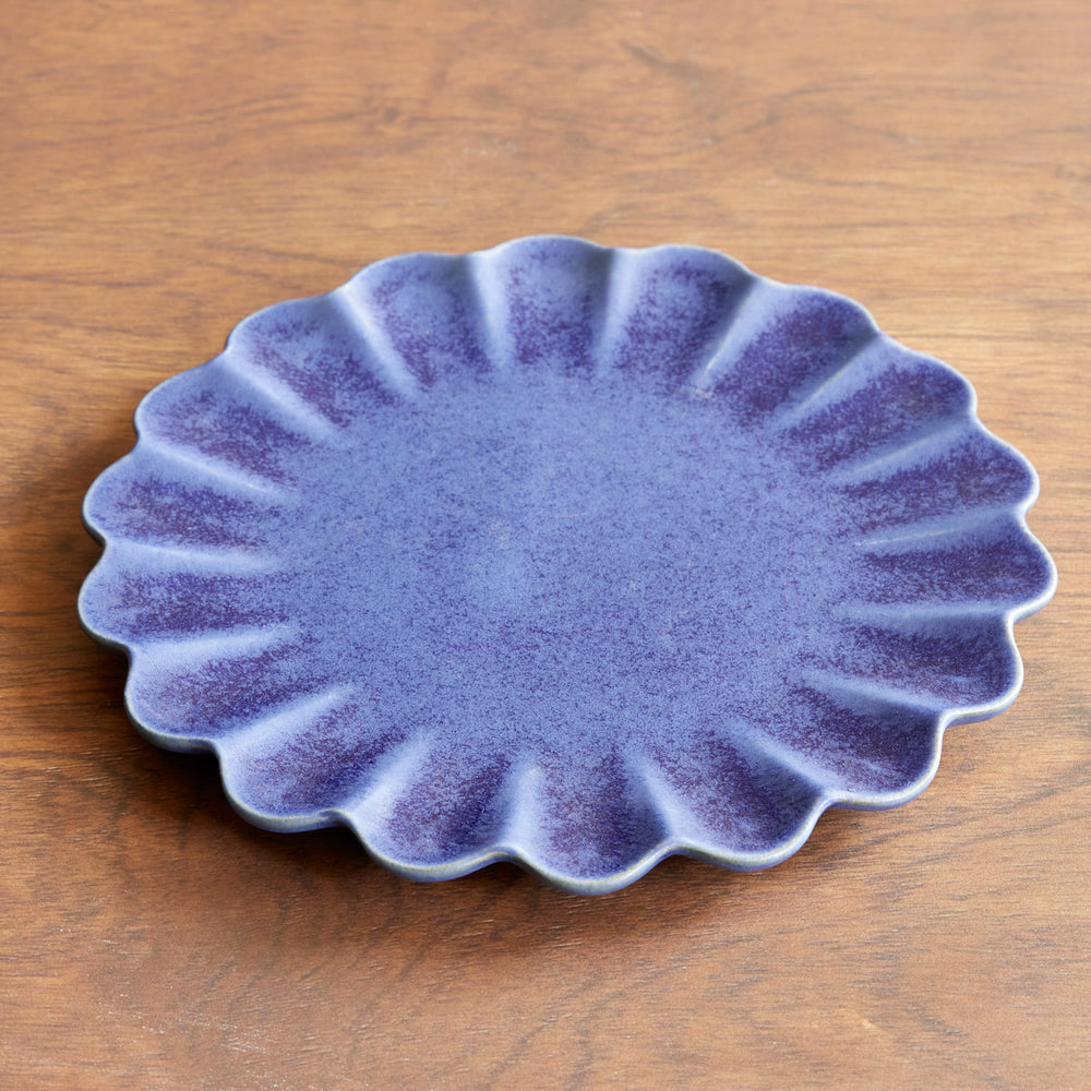 Lemieux et Cie Dhalia Stoneware Dinner Plate in blue ceramic with scalloped edges, shown on warm wood surface in soft natural light.