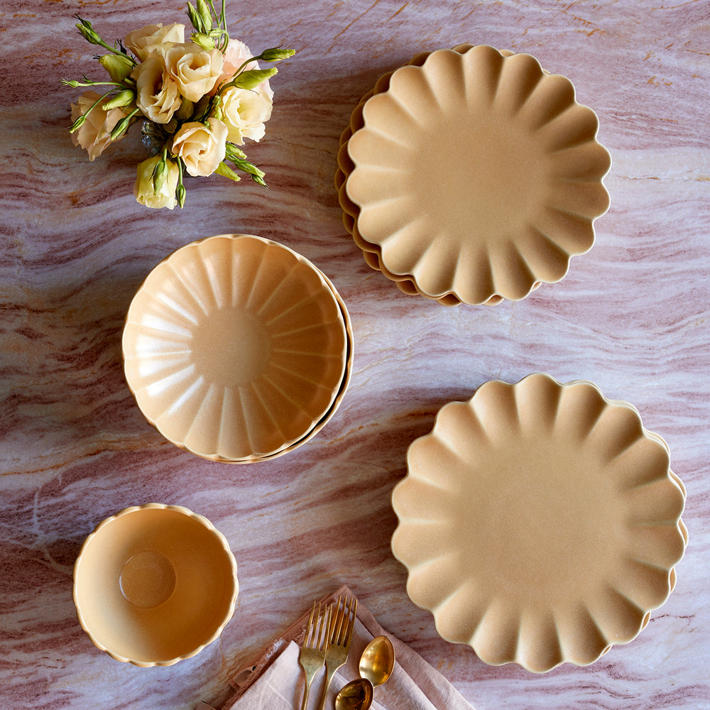 Lemieux et Cie Dhalia Stoneware dinnerware set in camel with scalloped edges, plates and bowls arranged on wood surface with pale roses and flatware.