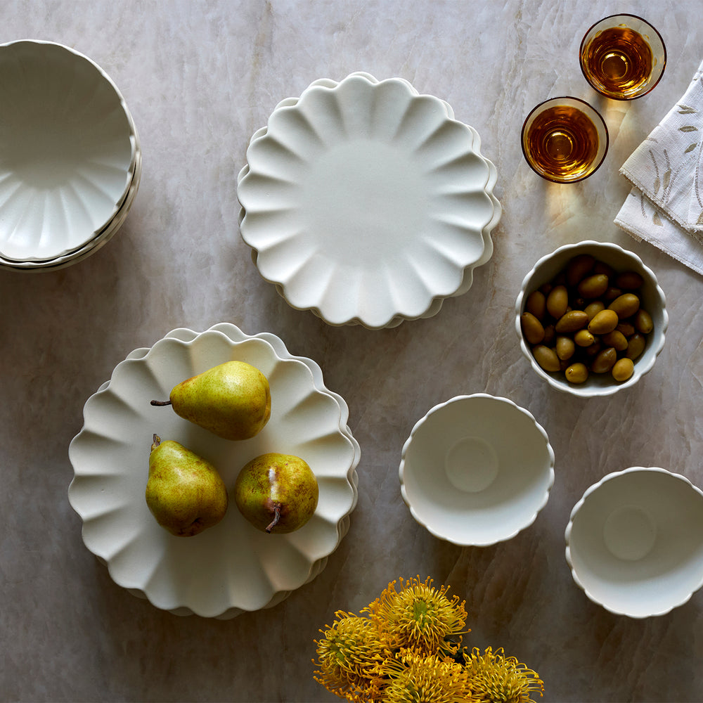 Lemieux et Cie Dhalia Stoneware dinnerware set in white, scalloped plates and bowls arranged on marble surface with pears, olives, amber glassware and linen napkin.