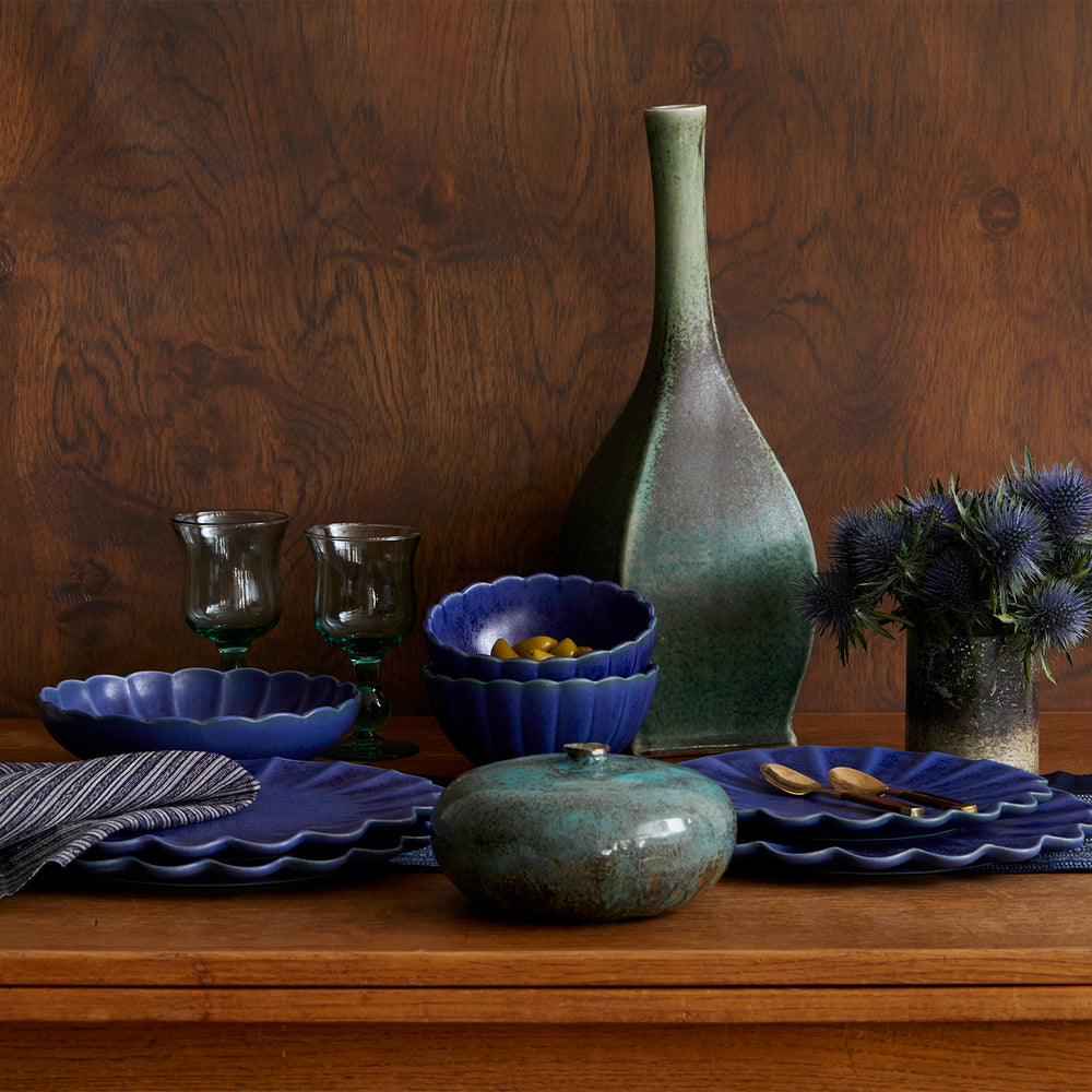 Lemieux et Cie Dhalia Stoneware dinnerware set in blue, scalloped plates and bowls styled with green ceramic vase and glassware on wood table against wood-paneled wall.