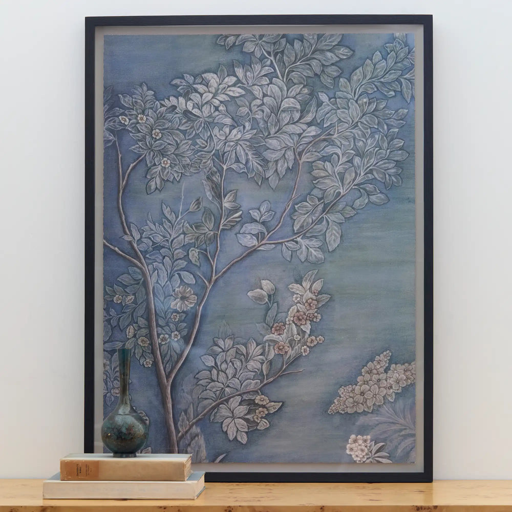 Lemieux et Cie Émeraude Un framed fine art print, botanical motif in blue-grey tones with silver branches and soft blossoms, displayed on wood surface with stacked books.