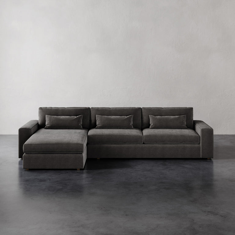 Lemieux et Cie Lacroix Left Chaise Sectional in charcoal upholstery with bolster pillows, modular sofa shown on dark concrete floors against white walls.