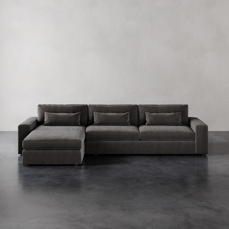 Lemieux et Cie Lacroix Left Chaise Sectional in charcoal upholstery with bolster pillows, modular sofa shown on dark concrete floors against white walls.