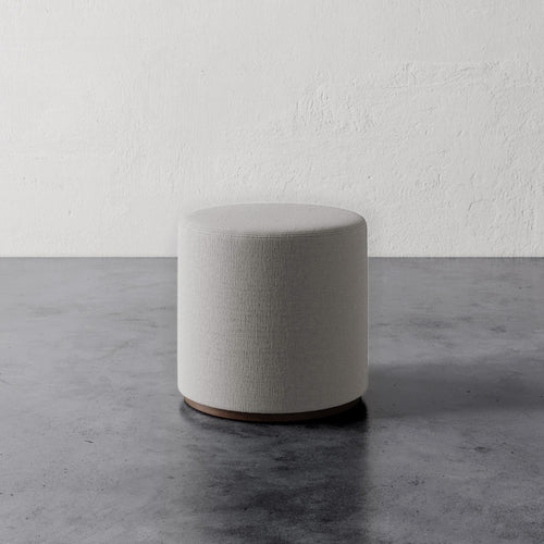 Lemieux et Cie Sophie Round Ottoman in sage upholstery with rounded cylindrical form, shown on concrete floor against textured plaster wall.