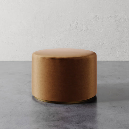 Lemieux et Cie Sophie Round Ottoman in warm caramel leather, low cylindrical form on concrete floor against textured plaster wall.