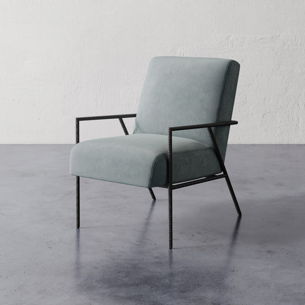 Lemieux et Cie Imane Lounge Chair, upholstered armchair with slim dark frame, shown on polished concrete floor against textured white plaster wall.