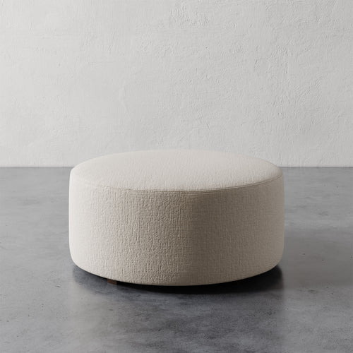 Lemieux et Cie Fabien 36" Round Ottoman in ivory textured upholstery, low-profile cylindrical ottoman shown on concrete floor against textured plaster walls.