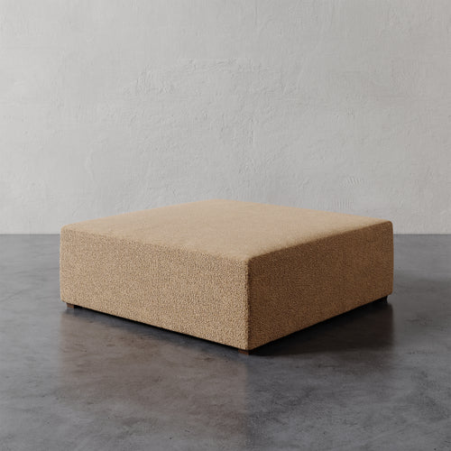 Lemieux et Cie Fabien 48" Square Ottoman in warm beige, low-profile upholstered ottoman on stone floor against textured plaster wall.