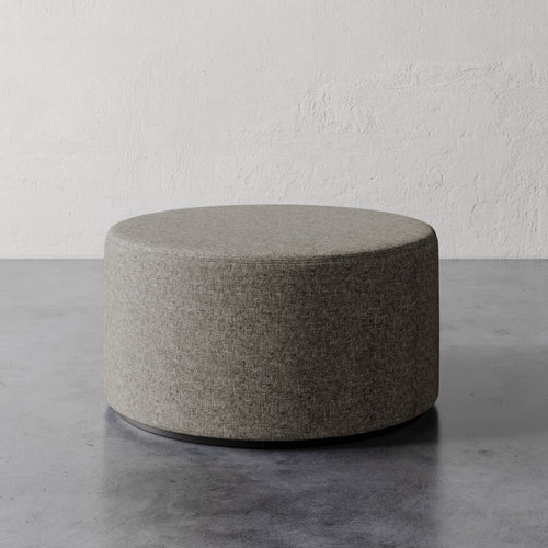 Lemieux et Cie Sophie Round 36" Ottoman in warm gray upholstery, low-profile ottoman on stone floor against textured plaster walls.