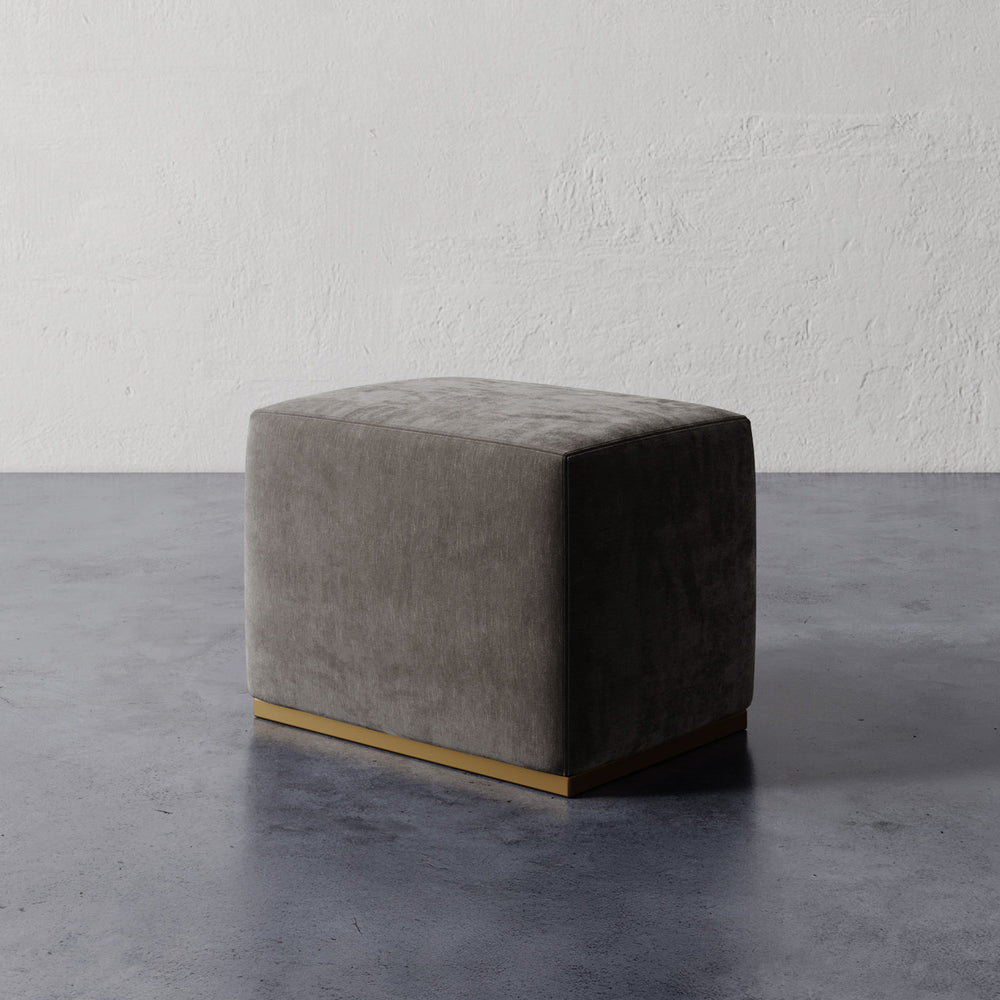 Lemieux et Cie Sophie Rectangular Ottoman with brass base, low-profile upholstered piece shown on polished concrete floor against textured plaster wall.
