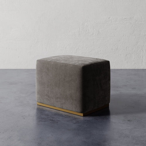 Lemieux et Cie Sophie Rectangular Ottoman with brass base, low-profile upholstered piece shown on polished concrete floor against textured plaster wall.