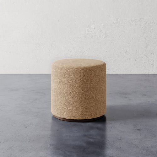 Lemieux et Cie Sophie Round Ottoman in warm bouclé, cylindrical upholstered ottoman with dark base, shown on concrete floor against textured plaster wall.