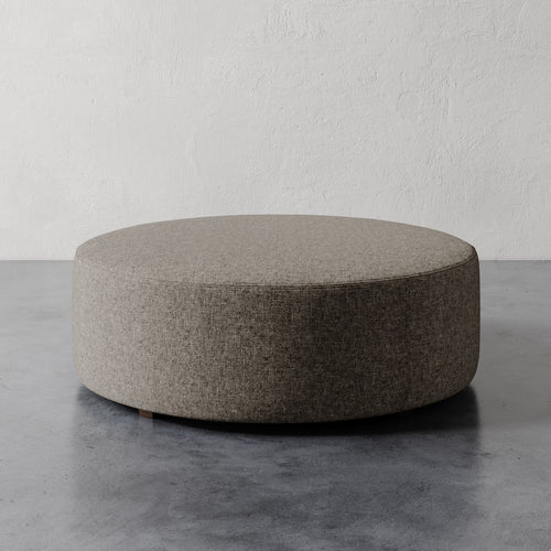 Lemieux et Cie Fabien 48" Round Ottoman in warm taupe upholstery, low-profile circular ottoman on concrete floor against textured plaster walls.