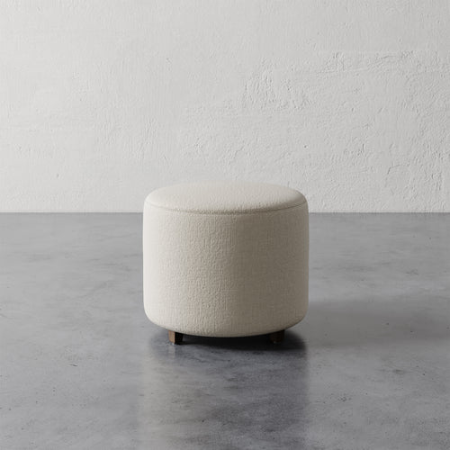 Lemieux et Cie Fabien 22" Round Ottoman in ivory textured upholstery, low cylindrical ottoman on concrete floor against textured plaster walls.