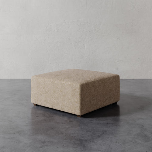 Lemieux et Cie Fabien 36" Square Ottoman in neutral upholstery, low-profile ottoman on dark concrete floor against textured plaster walls.