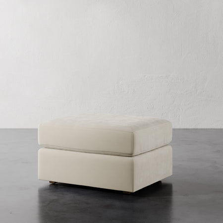 Lemieux et Cie Lacroix Ottoman, low-profile rectangular form in ivory upholstery, shown on dark concrete floor against textured white plaster wall.