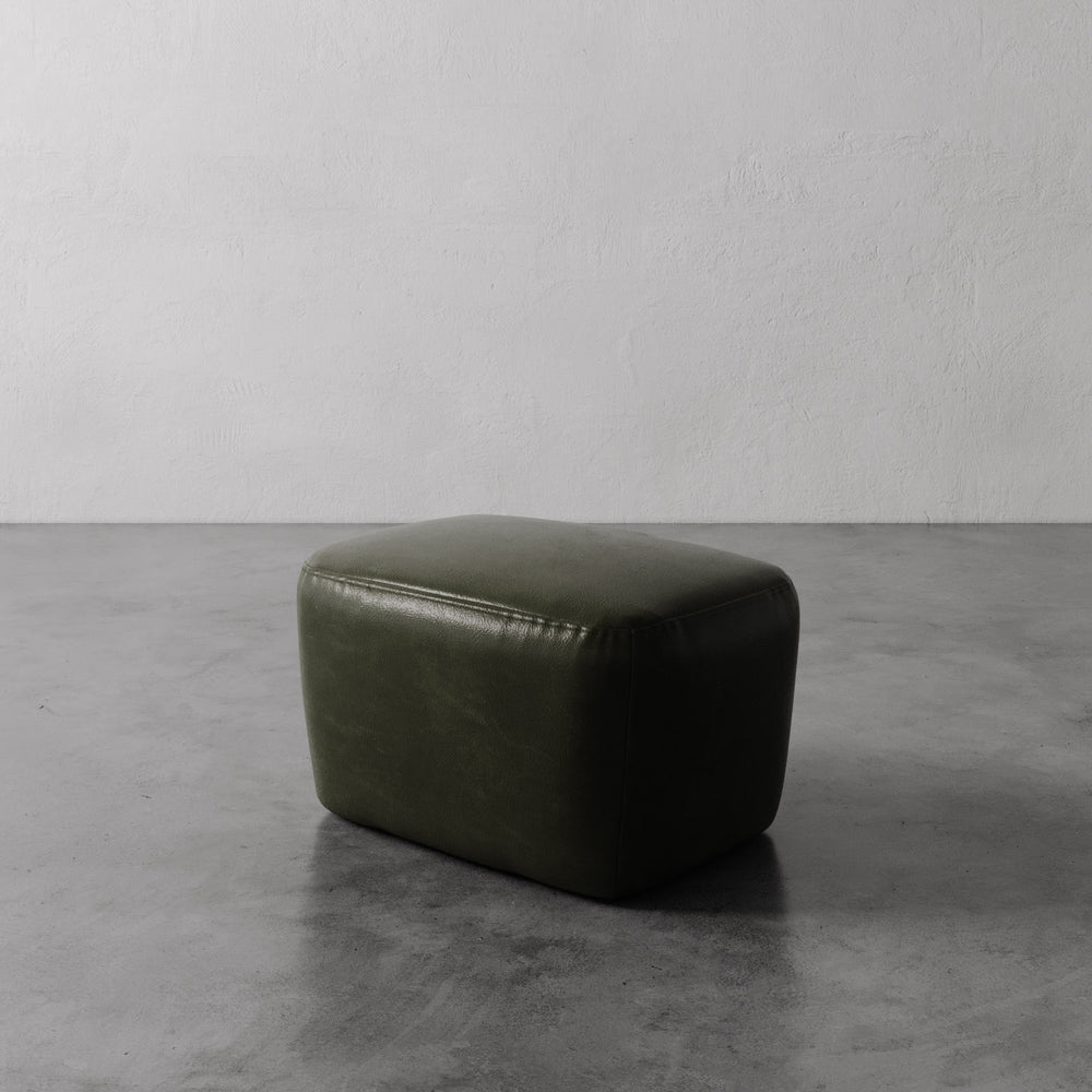 Lemieux et Cie Aurélie Leather Ottoman in olive green leather, low-profile ottoman with rounded form on concrete floor against textured plaster wall.