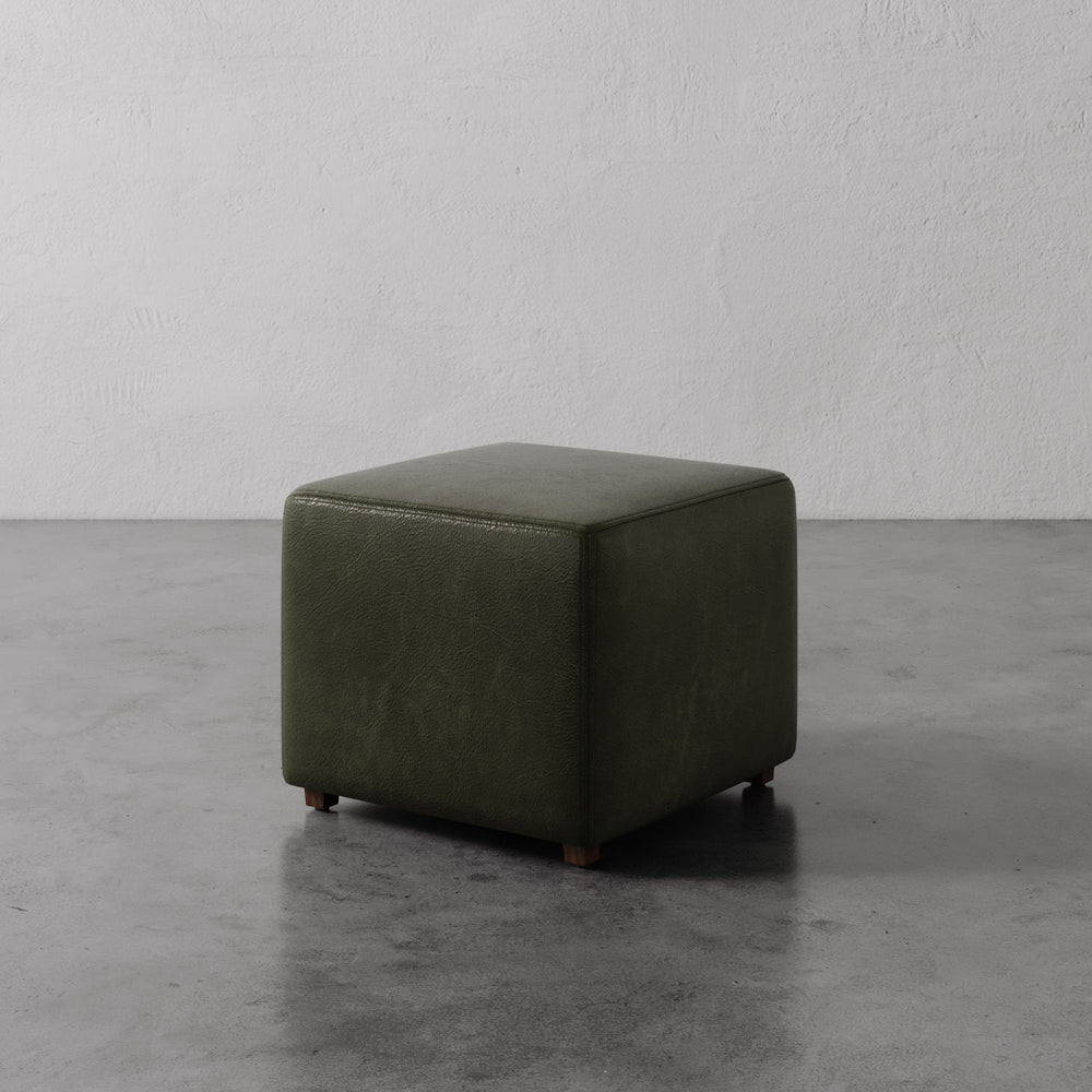 Lemieux et Cie Fabien Square Ottoman in olive green leather, low-profile cube form on turned legs, shown on concrete floor against textured plaster walls.