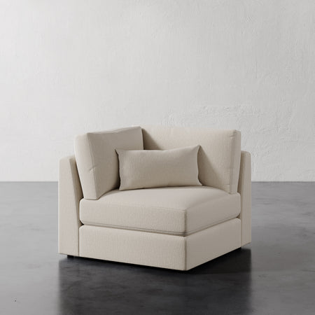 Lemieux et Cie Lacroix Corner Chair in neutral upholstery, modular sectional piece with deep cushions, shown on dark concrete floor against textured plaster walls.