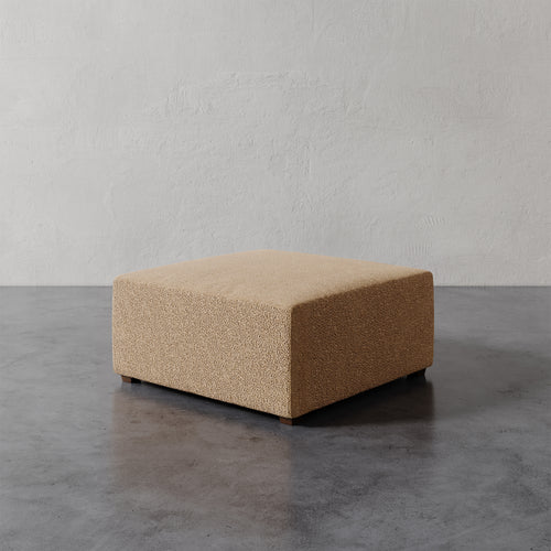 Lemieux et Cie Fabien 36" Square Ottoman in warm tan textured upholstery, low-profile geometric ottoman on concrete floor against neutral plaster wall.