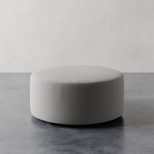 Lemieux et Cie Fabien 36" Round Ottoman, low-profile upholstered ottoman in neutral textured upholstery, shown on concrete floor against textured plaster wall.