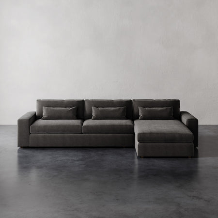 Lemieux et Cie Lacroix Right Chaise Sectional with bolster pillows, low-profile modular sofa in charcoal, on dark concrete floor against plaster walls.