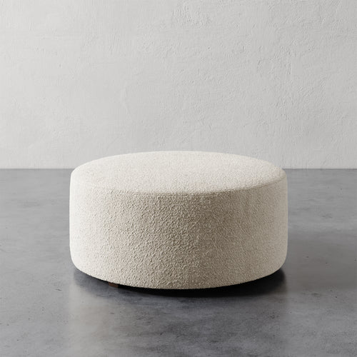 Lemieux et Cie Fabien 36" Round Ottoman in ivory bouclé, low-profile circular ottoman on polished concrete floor against textured plaster walls.